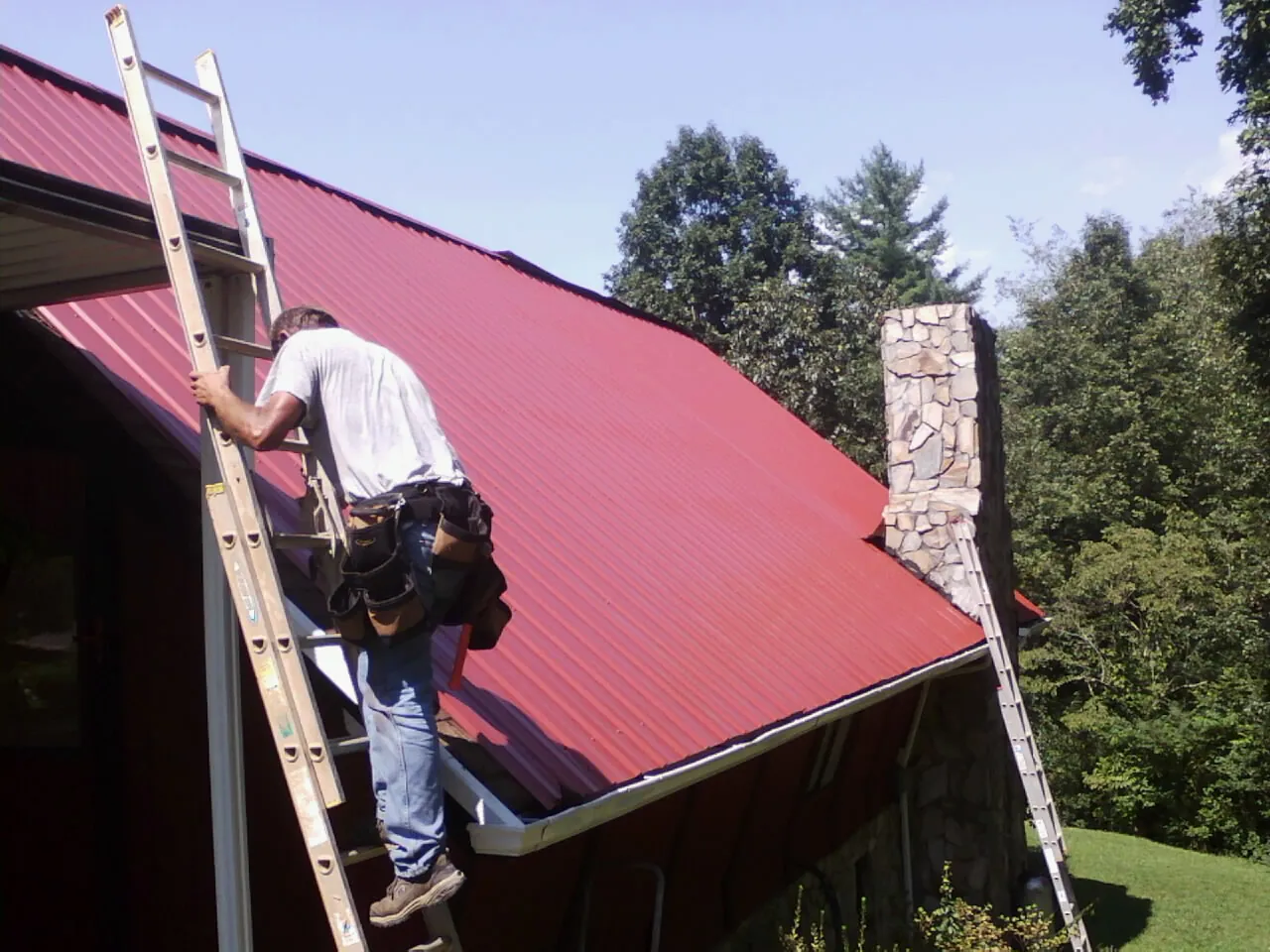Roofer on a ladder beside a red metal roof and stone chimney — Soffit & Fascia Repair by Roofing Brothers in Hot Sulphur Springs